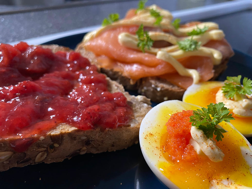 A close-up of an artisanal breakfast plate featuring seeded bread with strawberry jam, smoked salmon toast with mayonnaise, and soft-boiled eggs topped with roe and parsley.