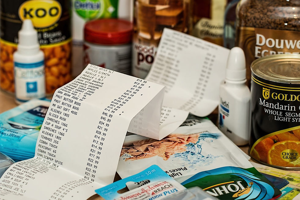 A long grocery receipt sits among various household items including canned goods, coffee, medicine, and toiletries, highlighting the cost of everyday essentials.