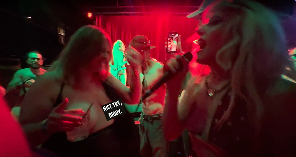 Lead singer with platinum blonde hair passionately sings into a microphone while interacting with an enthusiastic crowd at a live punk rock concert; red and green stage lighting sets an intense, high-energy atmosphere. A humorous sticker on a fan’s outfit reads “NICE TRY, DIDDY,” adding to the wild, unfiltered vibe.