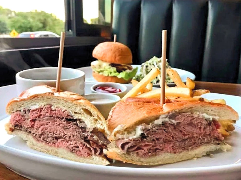 Close-up of a plated roast beef sandwich cut in half, stacked with thick slices of meat on a toasted bun and secured with wooden skewers. The sandwich is served with golden French fries and a small cup of dipping sauce. In the background, a cheeseburger with lettuce and tomato, a cup of ketchup, and a side of coleslaw are visible on a booth table by the window.
