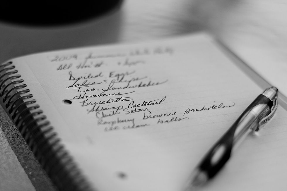 A black-and-white image of a handwritten food list in a spiral notebook with a pen resting on top, featuring items like deviled eggs and shrimp cocktail.