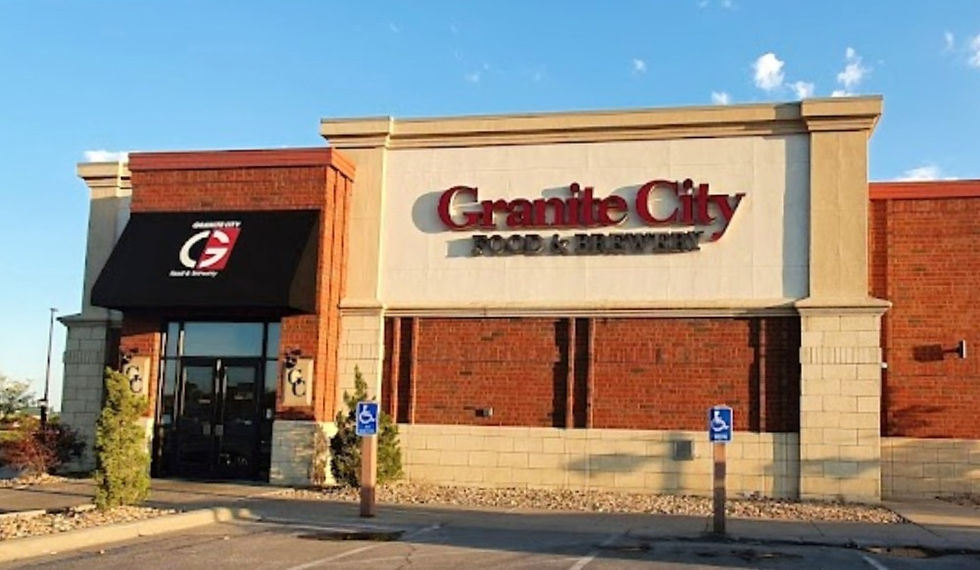 Exterior of a Granite City Food & Brewery restaurant building featuring a mix of red brick and light-colored stone facade. The main sign is prominently displayed in bold red and black lettering above the entrance. A black awning with the Granite City logo covers a side door. The parking area in front includes several designated handicap spaces, and the scene is lit by golden hour sunlight under a clear blue sky.