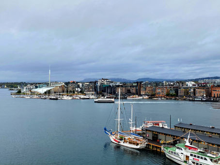 Scenic view of a harbor in Oslo, Norway, featuring sailboats and yachts docked near a modern waterfront with city buildings in the background under a cloudy sky.
