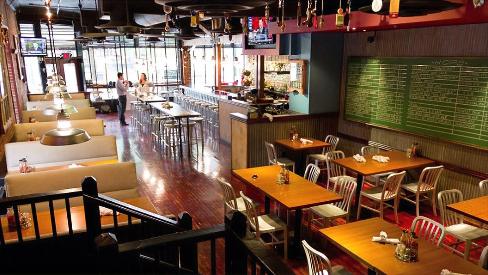Interior of a spacious, well-lit restaurant featuring a mix of booth seating and wooden tables with metal chairs. A long bar with high stools runs along the left side, and a large green chalkboard menu covers most of the right wall. Pendant lights hang over the booths, and several TVs are mounted above the bar. The polished wood floor and open layout create a warm, inviting atmosphere.