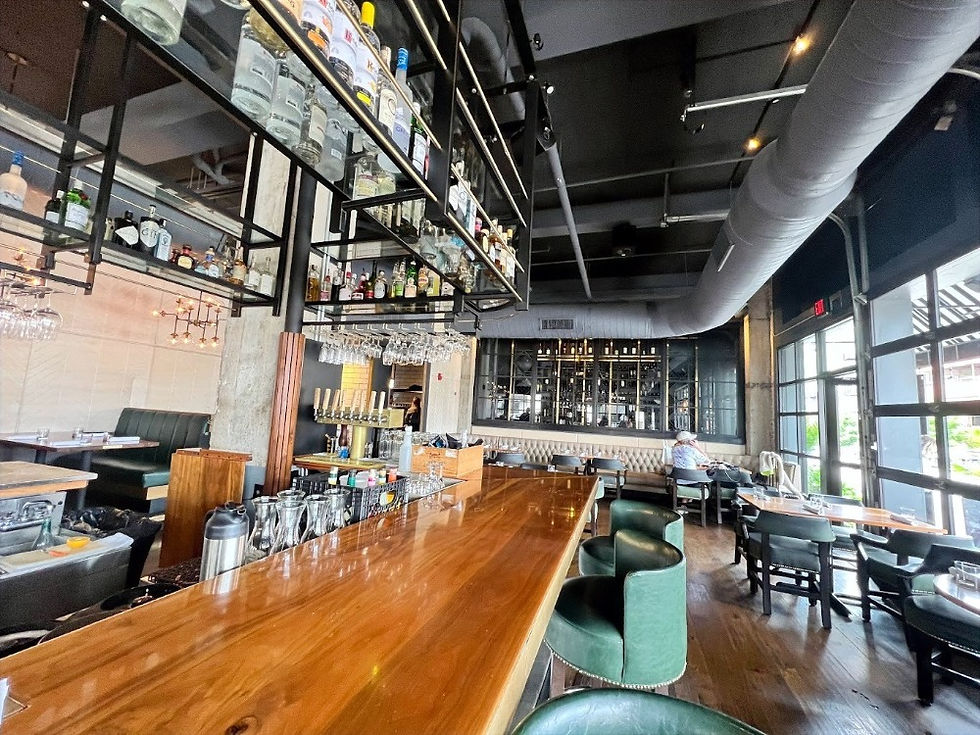 Interior of a modern, upscale bar and restaurant with a long polished wooden bar counter, green upholstered bar stools, and overhead shelves stocked with liquor bottles and hanging glassware. The space features dark exposed ceilings, an industrial-style air duct, and large windows letting in natural light. Booth seating and round tables are arranged throughout the open, stylish dining area.