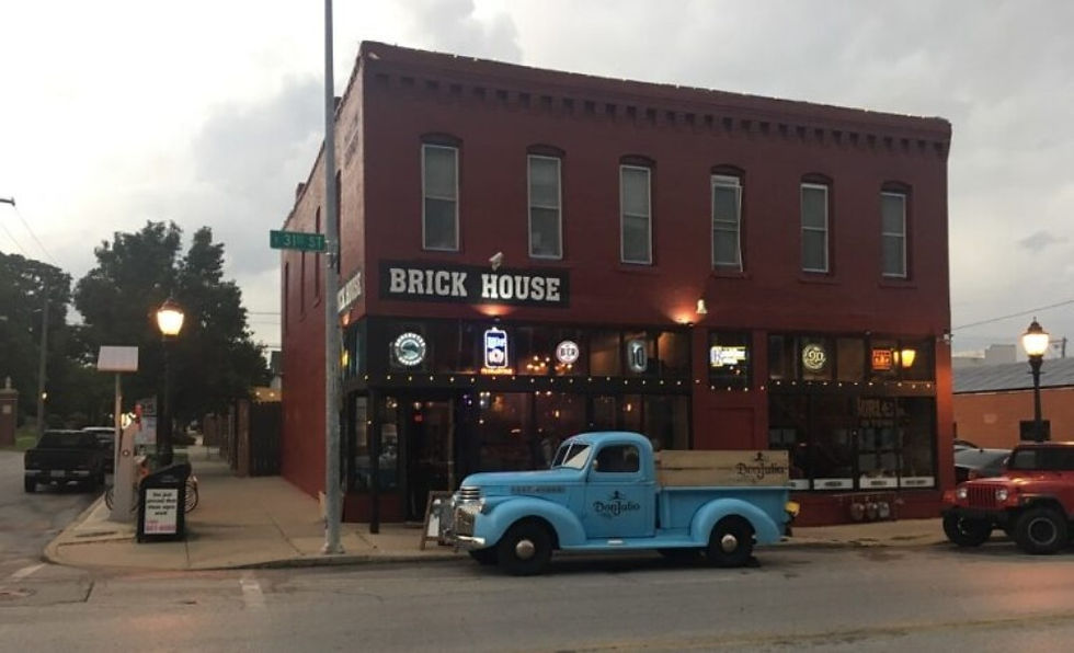 Street view of a red-brick two-story building housing a restaurant or bar named 'Brick House.' The building features several large windows with illuminated beer signs. A vintage light blue pickup truck with a wooden flatbed, labeled 'Doughboys,' is parked in front. The sky is overcast, and street lamps are lit as dusk settles over the quiet intersection.