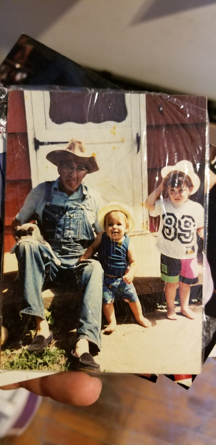 Grandfather wearing denim overalls and cowboy hat sitting with two young grandchildren and a small dog on a farm porch