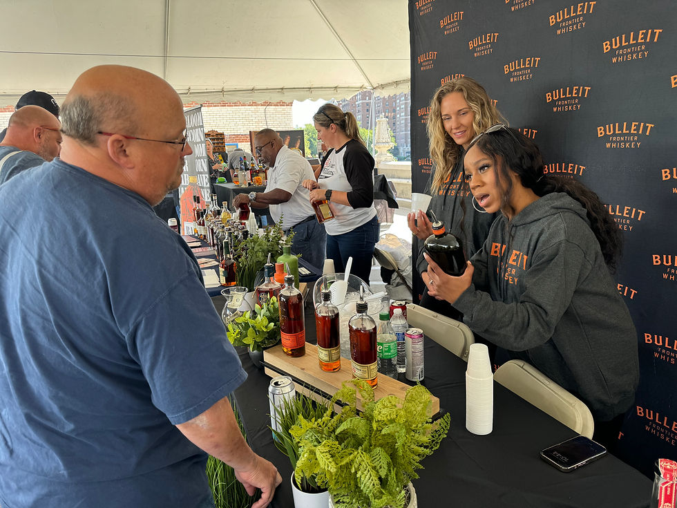 A woman at a Bulleit Whiskey booth enthusiastically describes a bottle to a visitor at an outdoor event, with whiskey bottles and greenery arranged on the table.