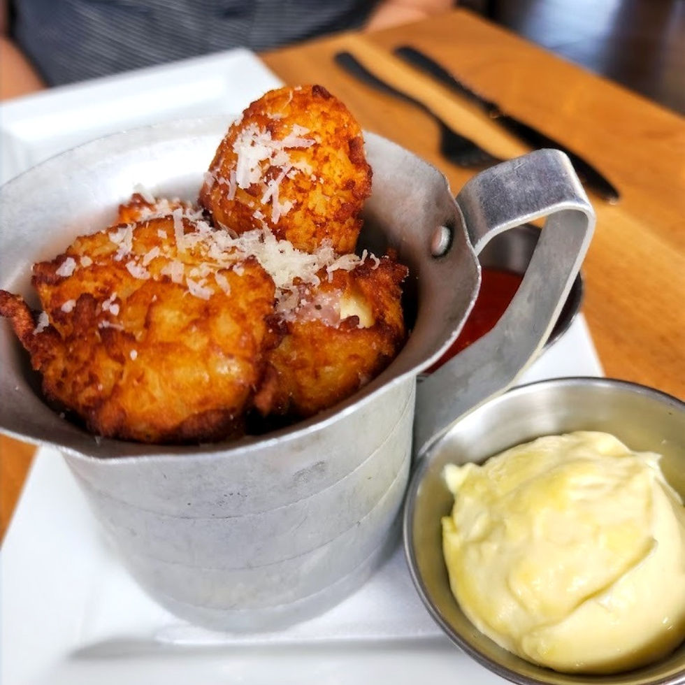 Close-up of a metal tin filled with golden brown, crispy tater tots topped with shredded cheese, served on a white plate. A metal ramekin of creamy dipping sauce is placed beside the tin. In the background, a person in a striped shirt sits at a wooden table with a knife and fork set nearby.