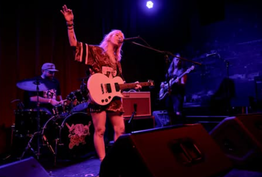 Indie rock band performing live under purple stage lighting, featuring a vocalist in a football jersey playing electric guitar, flanked by a bassist and drummer. The scene captures a moody, intimate venue vibe with strong stage presence and expressive performance energy.