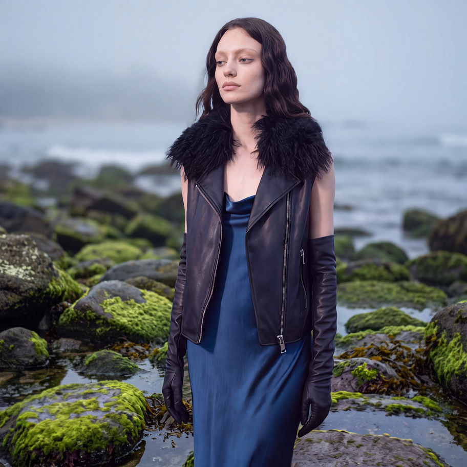 Model by the ocean wearing a black leather vest with a Mongolian fur collar, blue dress, and black opera gloves.