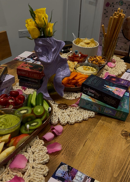 A table is laid with food, a goddess statue with yellow roses in a vase, tarot and oracle cards and candles