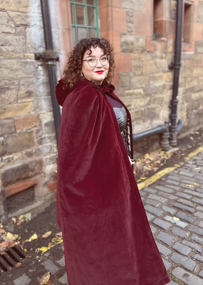 an adult woman stands on a cobbled street and looks back over her shoulder at the camera, she is wearing a burgundy cloak