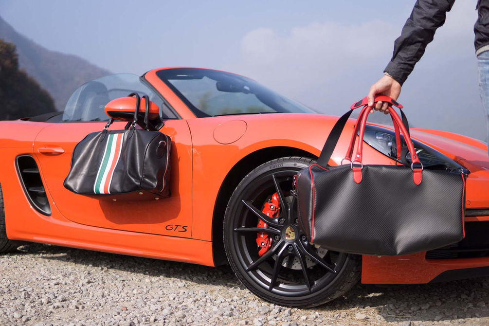 Two duffel bags by an orange sports car; black and carbon with red. 