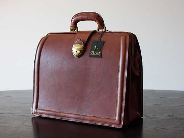 Brown leather bag with brass lock and a tag labeled "GLAM" on a dark wooden table. Clean, minimalist background.