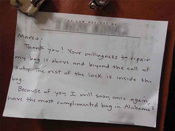Note with handwritten text attached to a brown bag. It thanks Marco for repairing a bag lock, expressing gratitude and humor.
