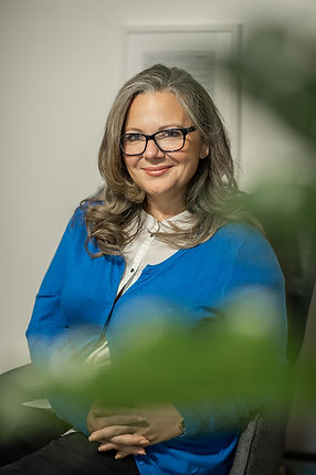 Mariangela Carta mit Brille und weisse Bluse und blaue Strickjacke sitzt.