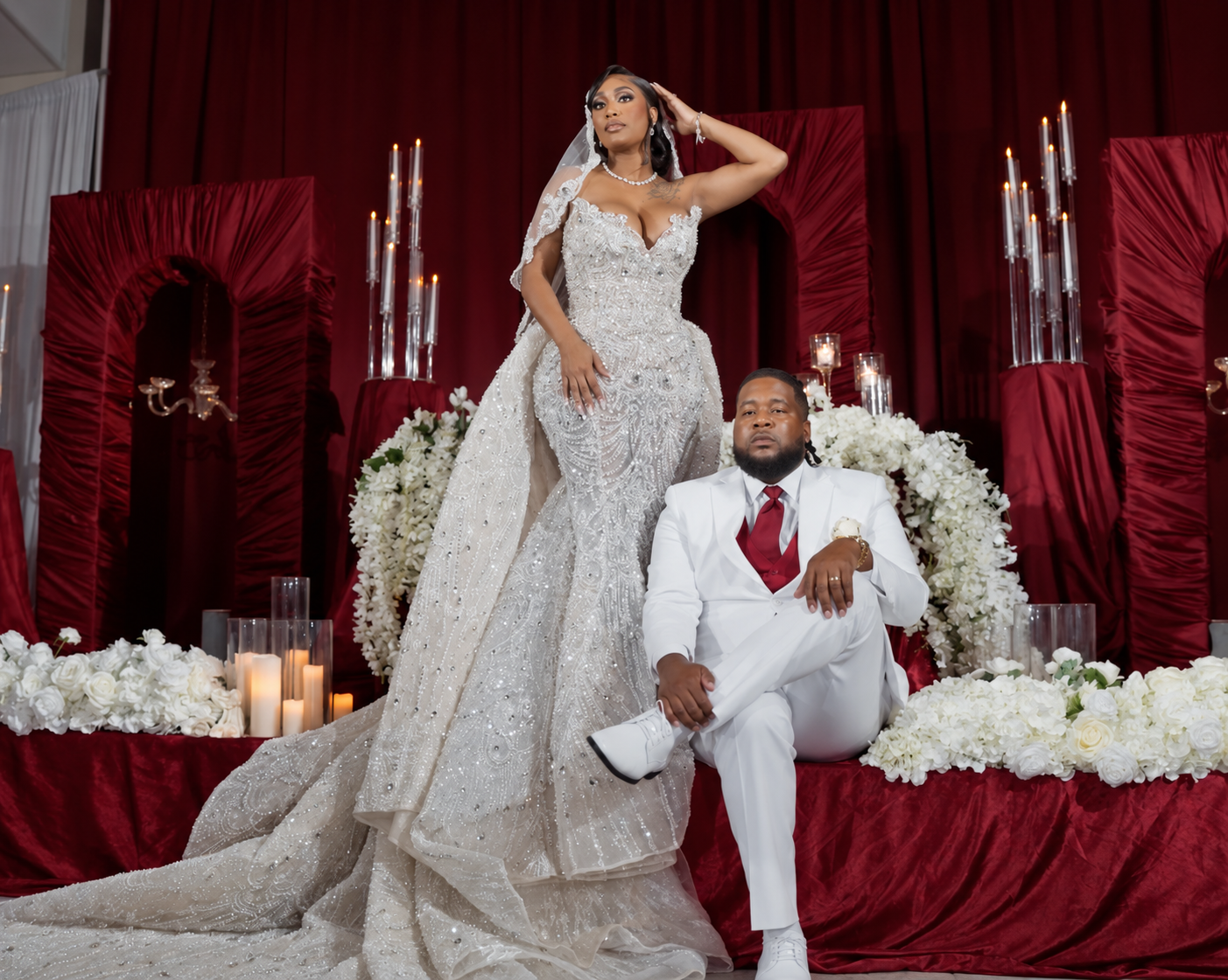 Bride standing beside her seated groom against burgundy drapes and white flowers