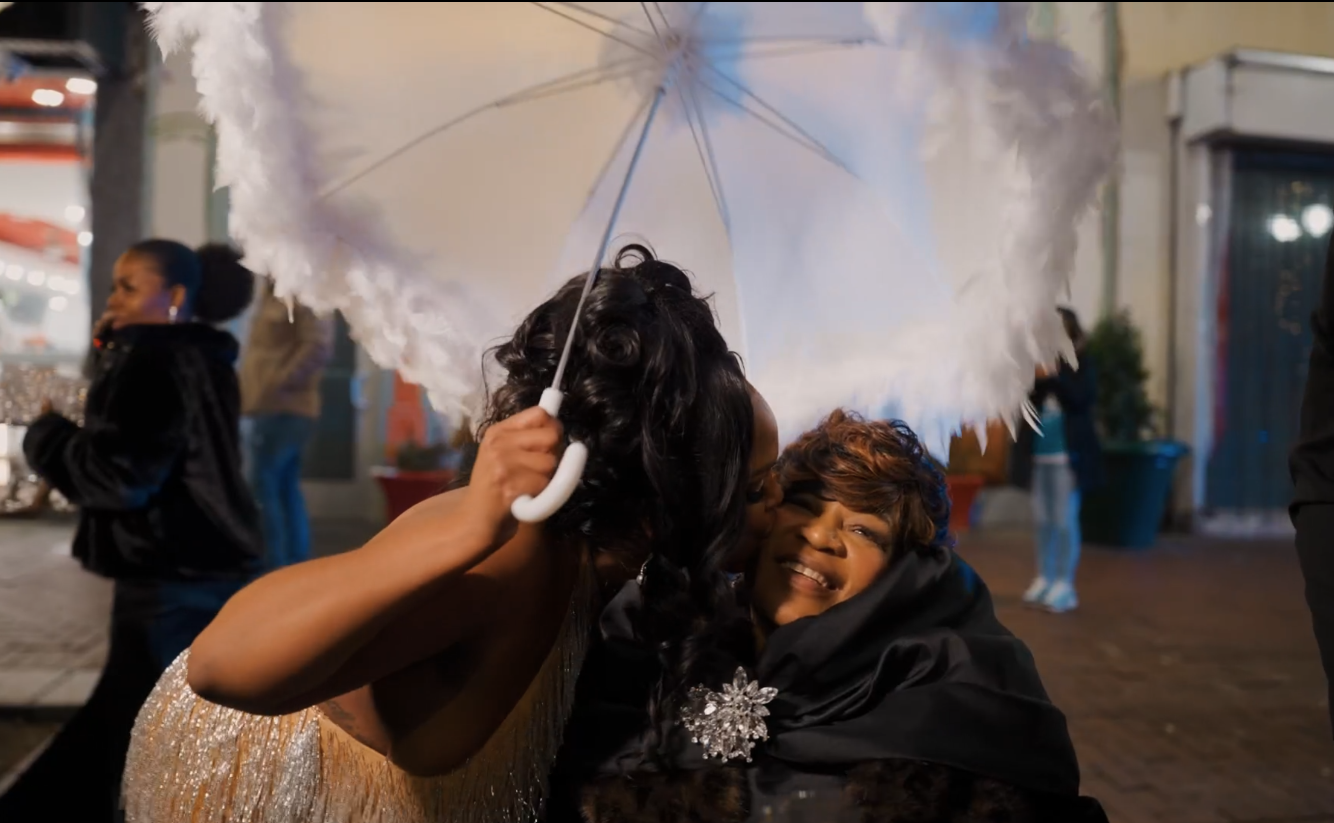 Bride sharing an affectionate moment with a smiling guest beneath a white feather-trimmed umbrella