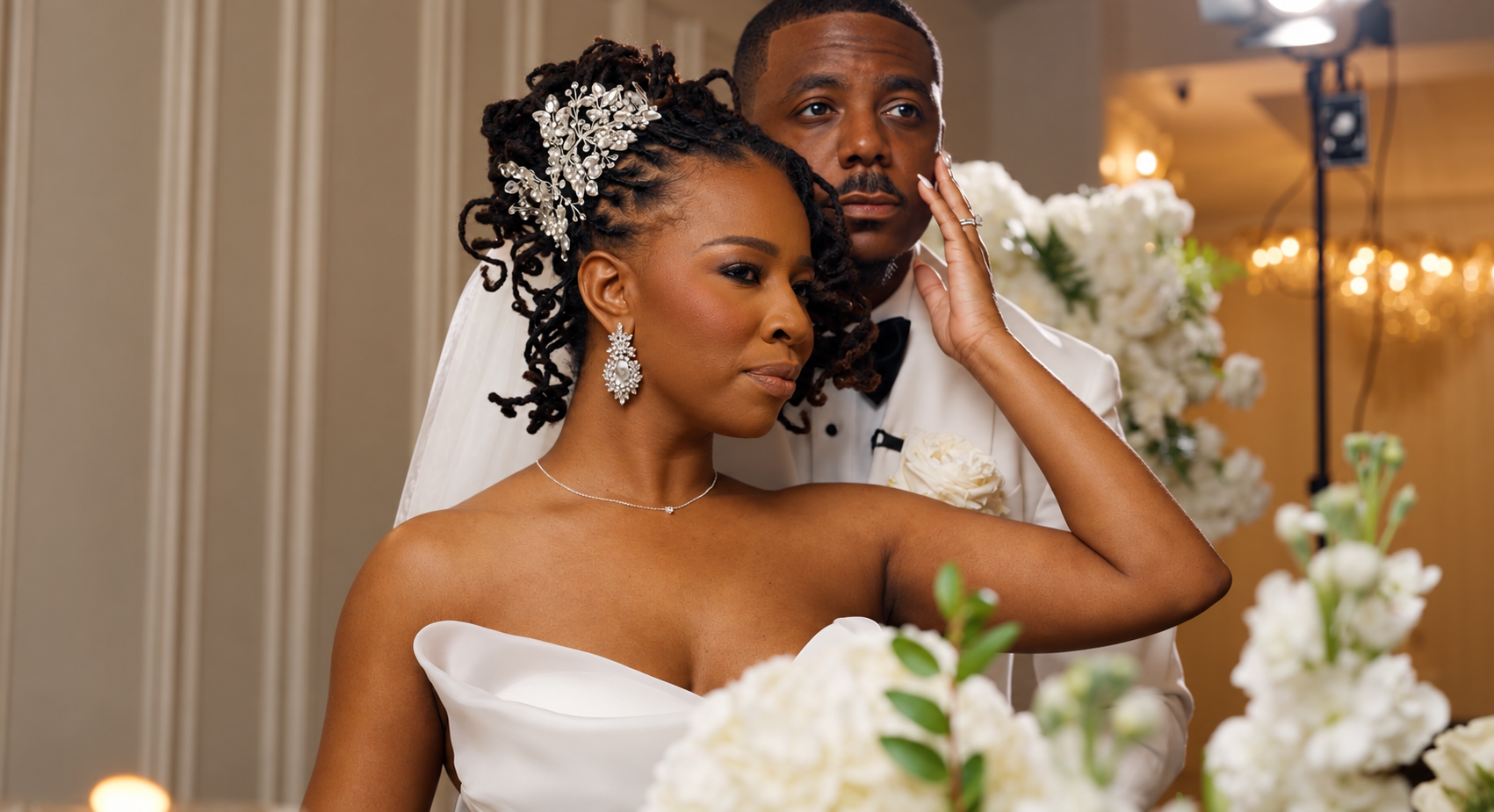 Bride reaching back to touch her groom’s cheek beside white wedding flowers