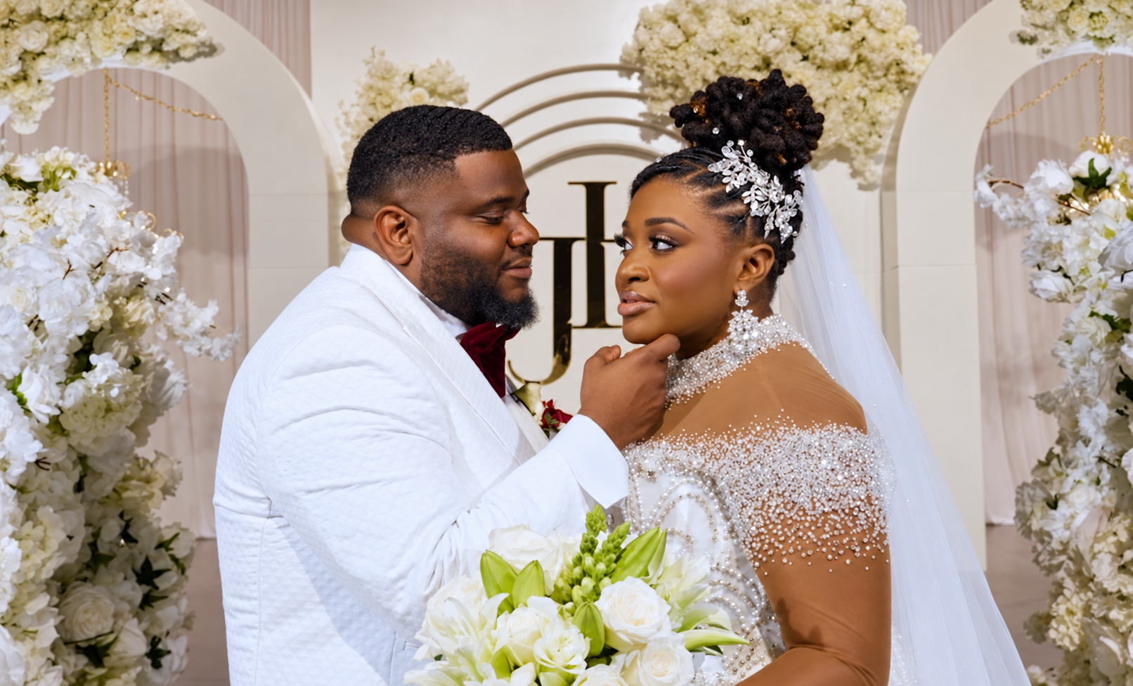 Groom gently touching his bride’s chin in front of a white floral backdrop