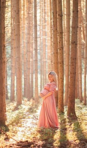 Schwangere Frau im rosa Kleid steht im sonnendurchfluteten Wald, hält Bauch.