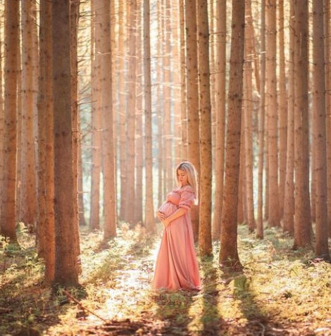 Schwangere Frau im langen rosa Kleid steht im sonnigen Wald.