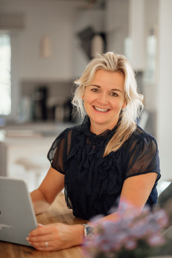 Lächelnde blonde Frau schaut in die Kamera, arbeitet am Laptop zu Hause.