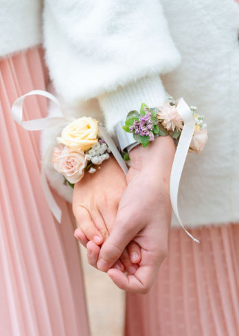 Zwei Hände halten sich, tragen zarte florale Handgelenks-Corsagen mit weißen Bändern.