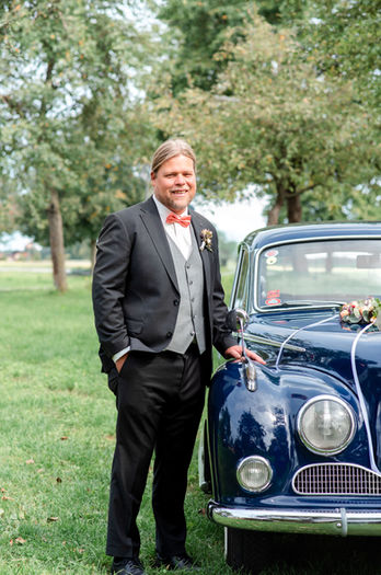 Bräutigam im Anzug lächelt neben blauem Oldtimer auf grüner Wiese.