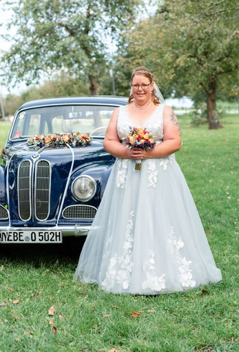 Frau im Brautkleid hält Blumenstrauß vor blauem Oldtimer WEBE O 502H.