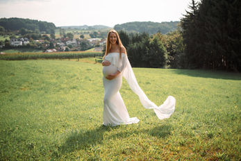 Schwangere Frau im weißen Kleid steht lächelnd in sonniger grüner Wiese.