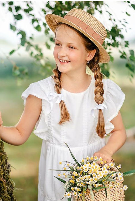 Lächelndes Mädchen in Strohhut, weißem Kleid hält Blumenkorb neben Baum.