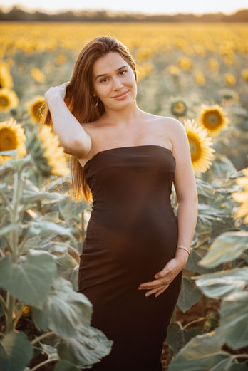 Schwangere Frau im schwarzen Kleid, lächelnd in Sonnenblumenfeld bei Sonnenuntergang.