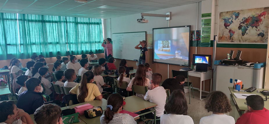 Photograph of a dark classroom, with closed curtains. Irene and a teacher are looking towards the back of the classroom through their hands (curved like a telescope). The room is filled with approximately 30 children all facing the demonstrators. Behind the demonstrators there is a white board and a screen displaying a large telescope.