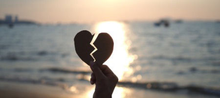 A hand holding up a paper broken heart out over the ocean against a glistening sunset