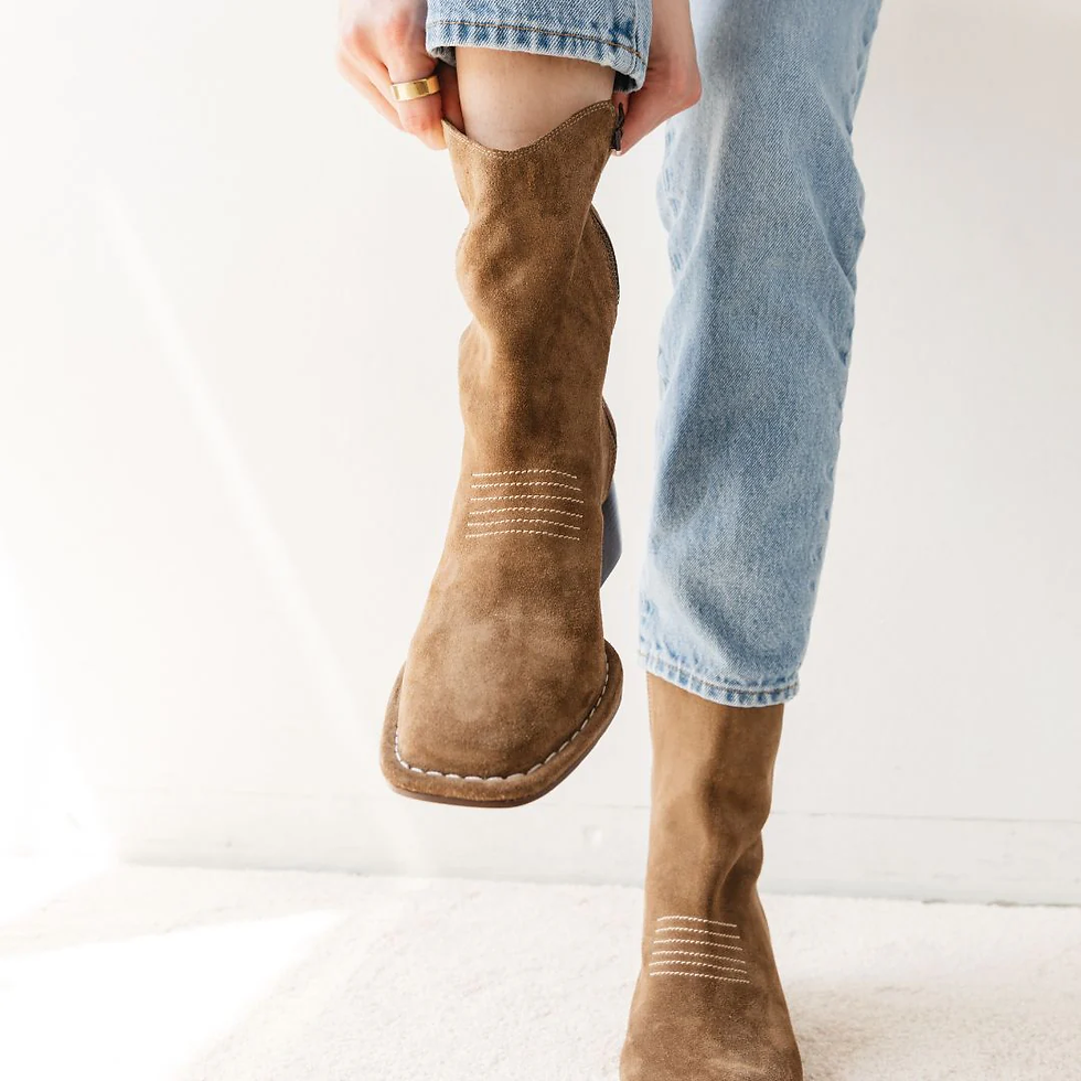 Kvinde tager på brune støvler, iført jeans, foran en hvid væg.