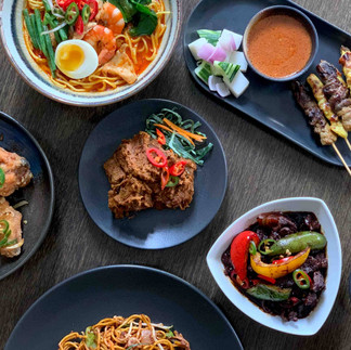 A sumptuous spread of assorted asian dishes on a wooden table, featuring noodles, grilled skewers, a fried egg topping, and flavorful sauces.