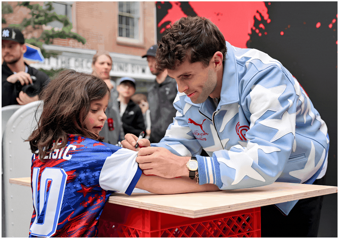 Christian Polisic signs young boy's 10 jersey at a table during an outdoor event, with onlookers and a red-black backdrop.
