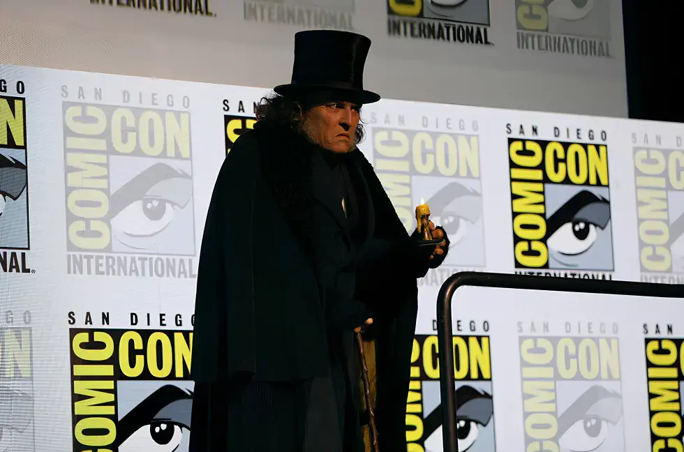 Johnny Depp as Ebenezer Scrooge in a black top hat and cloak stands at San Diego Comic-Con backdrop, holding a cane and small object, looking stern.