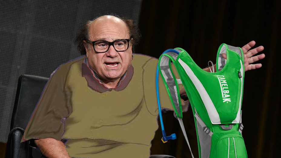 Danny DeVito dressed as Quasimodo holds up a green Camelbak backpack