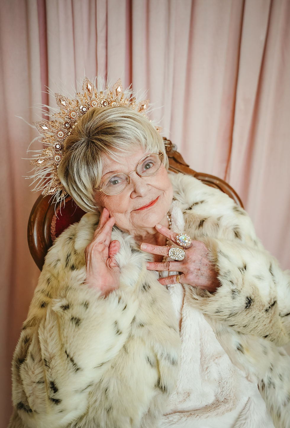 Elderly woman in a fur coat and jeweled crown poses smiling before pink curtains.