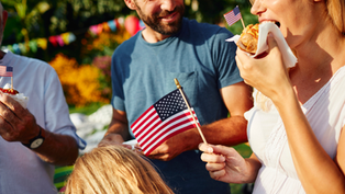 People eating hot dogs with small U.S. flags at a sunny outdoor party, smiling and chatting.