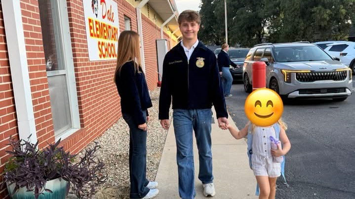 Smiling teen in FFA jacket walks with a little girl outside Dee Elementary School, with a woman nearby and parked cars.