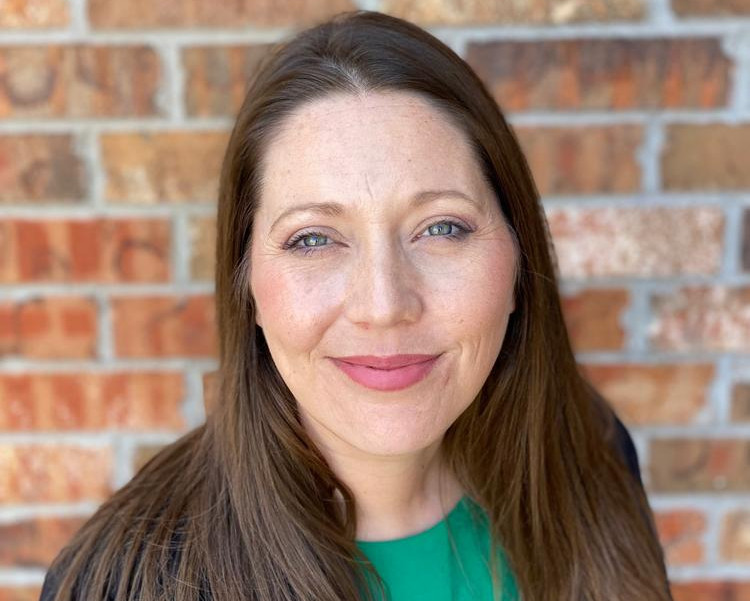 Smiling woman with brown hair in a green top and black blazer stands before a brick wall.