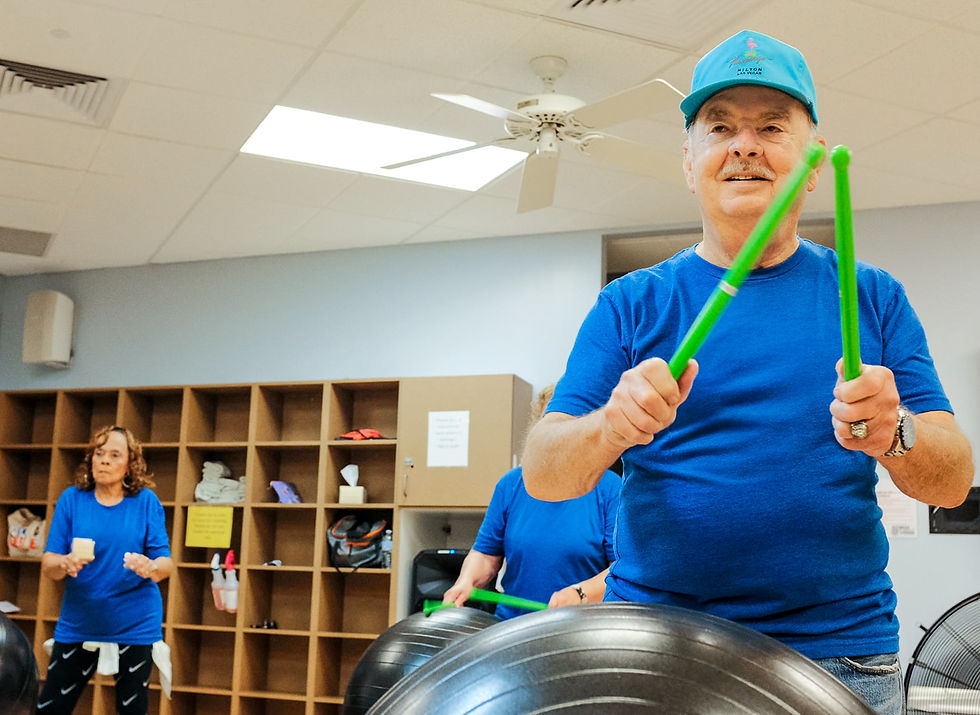 Older man in blue shirt and cap plays green drumsticks on exercise balls in a bright studio at Shawnee Recreation Center in Pottawatomie County, Oklahoma, with two women behind him.