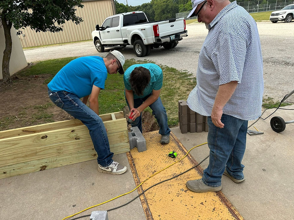 Three men build a wooden frame on a driveway, using cinder blocks and a drill, with trucks and a beige building in the background.