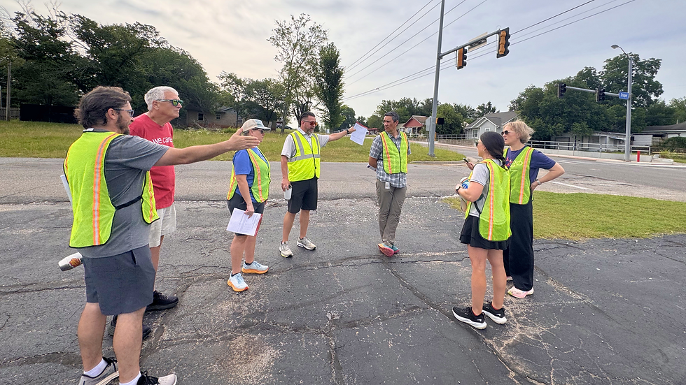 Shawnee walk audit participants in yellow safety vests discuss at an intersection. Green trees and traffic lights in the background under a cloudy sky.