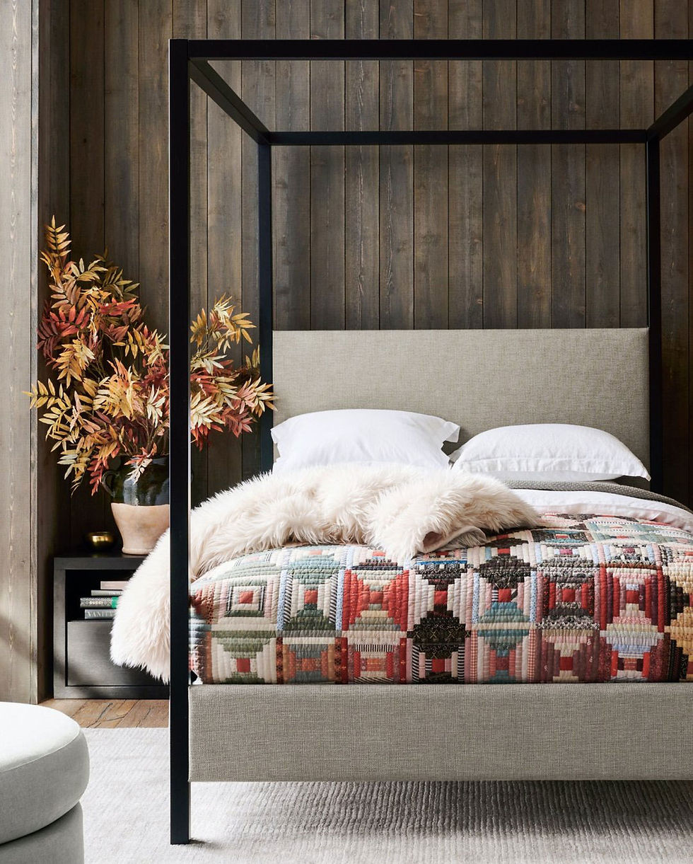 Cozy bedroom with black four-poster bed, patchwork quilt, white pillows, fur throw, and autumn foliage in a vase against wood walls.