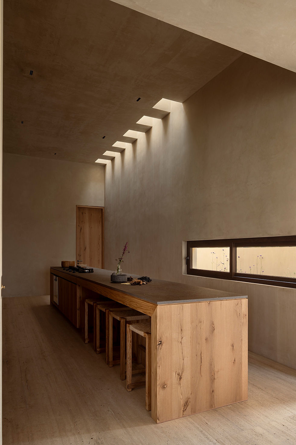 Minimalist beige dining room with a long wooden island, stools, skylight openings, and a narrow window with flowers.